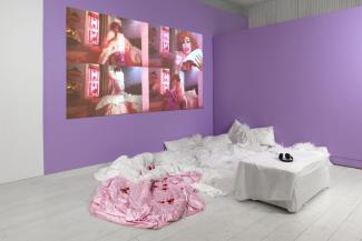 Installation view of feather boas, a video projection, and white seating installed in a purple corner of the gallery