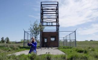 a video still of artist Alana Bartol kneeling on the grass in front of a fenced off orphan well
