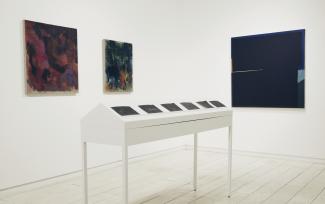 installation view of three dark paintings and a long white table