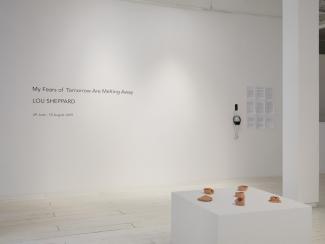Installation view of the exhibition title on a wall, with a plinth displaying ceramics in the foreground