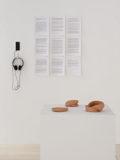 Installation view featuring a grid of nine documents on the wall, and an iphone with headphones installed to its left, with a plinth displaying unglazed ceramic forms in the foreground.