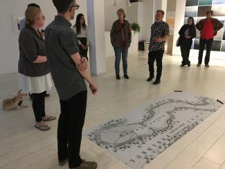 Artist Alex Grünenfelder stands in Access Gallery in front of a large black and white printed map laid on the floor with two train spikes on its corners. A small crowd of about 8 people, and one small dog, look on while Alex conducts his performance piece.