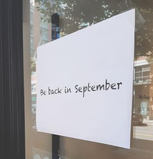 A piece of paper taped to the front door of Access Gallery stating "Be back in September".