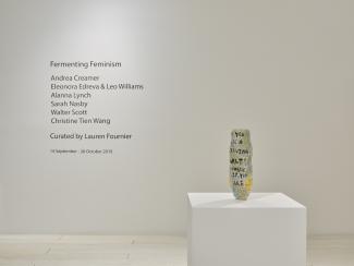 A ceramic vase on a plinth, with artists names on the wall behind
