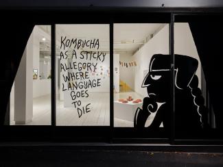 View into the lit gallery at night, through a window with a figure and letting in black vinyl "Kombucha is a sticky allegory where language goes to die"