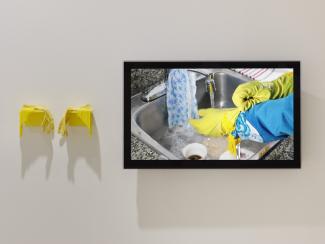 Image of a white wall on which installed a pair of beaded yellow rubber gloves and a flatscreen displaying the footage of two hands washing dishes in these gloves. 
