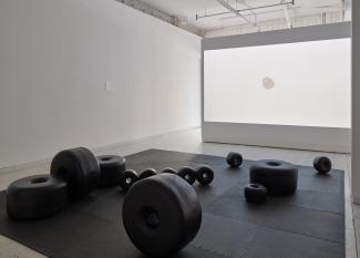 Installation image of large black sculptural beads scattered across a floor mat, with a video of a white beads accumulating in a circle on a white background, projected on the wall behind the sculptures.