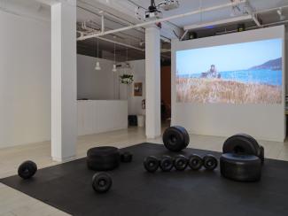 Installation image of large black sculptural beads scattered across a floor mat, with a video of a Newfoundland landscape projected on a wall behind the sculptures.