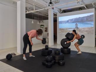 Installation image of Katie (in pink shirt and black denim pants) and Dana (in orange shirt and denim shorts) playing with large black sculptural beads scattered across a floor mat, with a video of a Newfoundland landscape projected on a wall behind the sculptures.