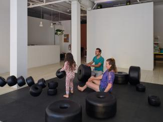 Installation image of a father in teal, mother in purple, and child in cherry print pyjamas playing amongst large black sculptural beads scattered across a floor mat.