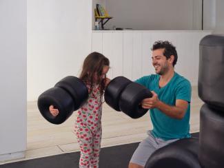 Installation image of a father in teal helping his child in cherry print pyjamas put large black sculptural beads on her arms like sleeves.
