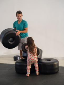Image of a father holding one of Carrie Allison's BIG BEADZ, sticking his tongue out in concentration, with his long-haired child in cherry-print pyjamas looking up at him, away from the camera, with two smaller BIG BEADZ worn on her arms.