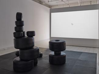 Installation image of large black sculptural beads arranged in towers on a floor mat, with a video of a white beads accumulating in a circle on a white background, projected on the wall behind the sculptures.