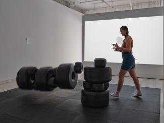 Installation image of Dana (earring orange and denim) pushing a tower of black sculptural beads and letting them tumble across a floor mat, with a video of a white beads accumulating in a circle on a white background, projected on the wall behind the sculptures.