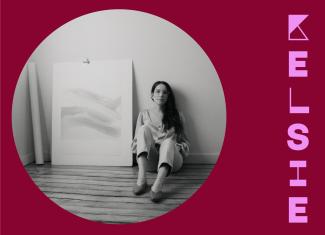 Circular image of black and white photo of Kelsie, sitting on the floor in her studio beside a large painting. The portrait is placed in a dark raspberry background with her stylized name in bright lavender to the right.