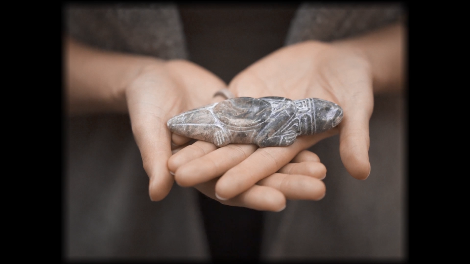 A still from Spirit Emulsion, showing artist Siku Allooloo's hands cradling a small reptile figurine carved from rock. The details are finely etched, with a small divot in the creature's back