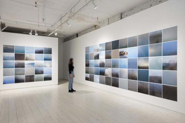 installation view of two large grids of photogs on adjacent walls, with a blurred figure observing