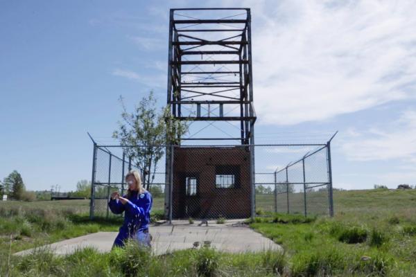 a video still of artist Alana Bartol kneeling on the grass in front of a fenced off orphan well
