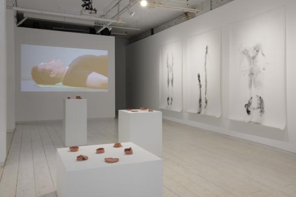 Installation view featuring three large drawings, a video of the artist lying on their back, and three plinths displaying unglazed ceramic forms.