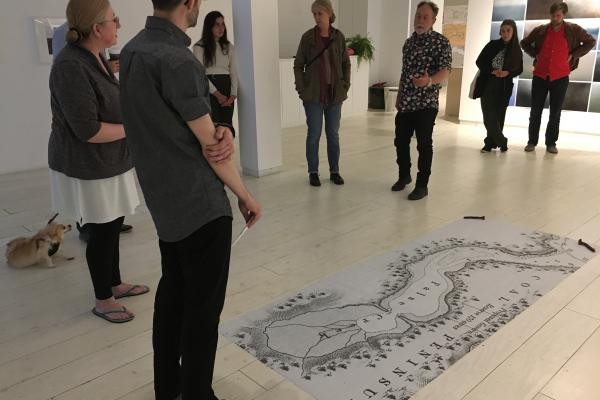 Artist Alex Grünenfelder stands in Access Gallery in front of a large black and white printed map laid on the floor with two train spikes on its corners. A small crowd of about 8 people, and one small dog, look on while Alex conducts his performance piece.