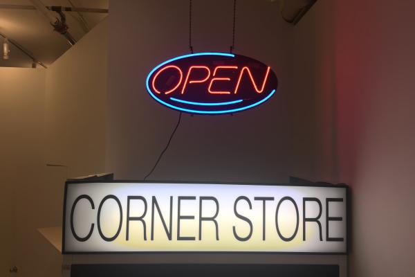 A neon-light 'Open' sign hangs above a drink fridge with a back-lit sign that reads 'CORNER STORE'. Photo taken in Access Gallery's PLOT space.
