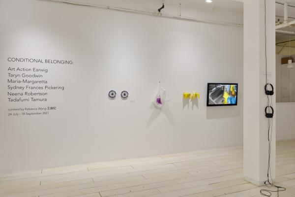 Image of a white wall and a white post in a gallery space. There are texts on the left side of the wall, next to which are two beaded blue and grey plates, a beaded translucent and fuchsia plastic bag, a pair of beaded yellow rubber gloves and a flatscreen displaying the footage of two hands washing dishes in these gloves. There are two sets of black headphones hung on the post to the right of the image.