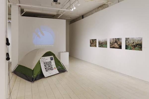 Image of a white gallery space with two perpendicular walls. There is a video projection showing two feet in rain on the end wall, a grey and green tent installation with a large QR code in front of the projection wall, and four colour photograph prints depicting human bodies in nature on the wall to the right.