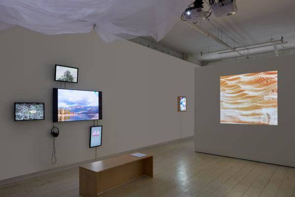 Three quarter installation view of gallery installation. On left wall is a four channel video installation of landscapes in vivid colour, on the right is a single projection of a carved blonde wood.
