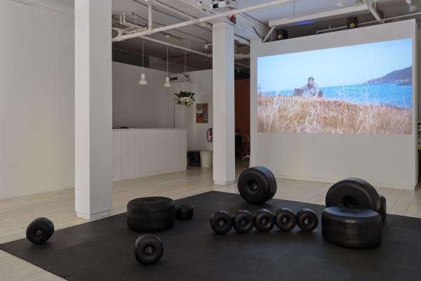 Installation image of large black sculptural beads scattered across a floor mat, with a video of a Newfoundland landscape projected on a wall behind the sculptures.