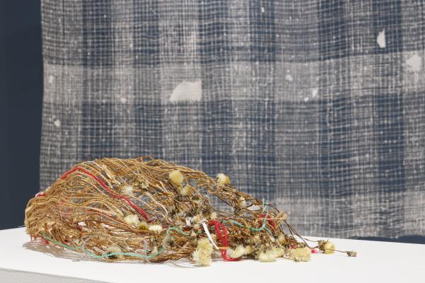 A woven basket made from dried flowers, grass, thread, and paper. It lies on its side on a white table in the foreground. In the background there is a white weaving in front of a blue wall. 