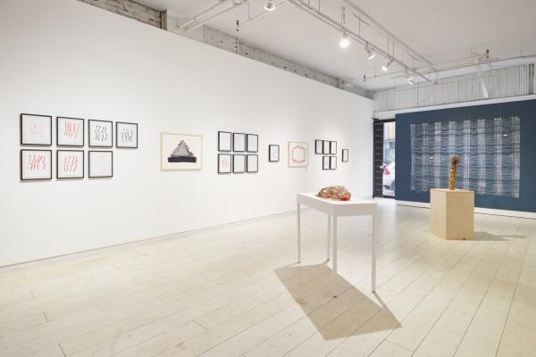 A gallery view of the exhibition. In the foreground a woven basket sits on a white table. Behind that a bundle of dried flowers rests upright on a wooden plinth. On the left wall there are many framed works including weavings and handmade paper. The far wall is painted blue, and there is a white weaving hanging in front of it. 