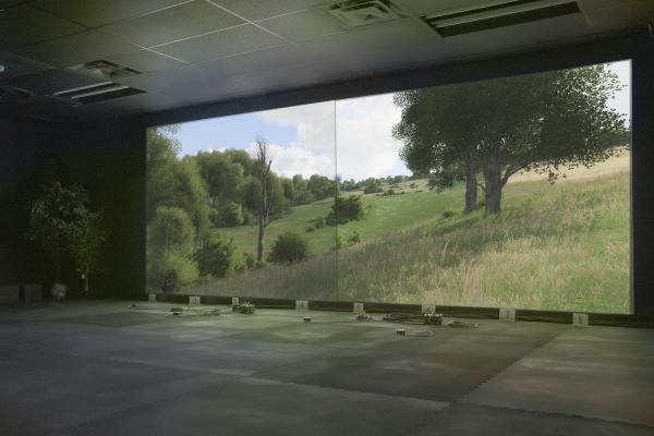 Steven Cottingham, Virtual Range, 2025. The image shows a tranquil, digitally simulated landscape with trees and grass clearings. The image is projected on the wall of a dimly lit shooting range at a military base in Petewawa, Ontario. 