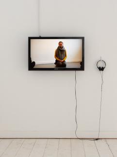 Installation view featuring of a wall mounted TV monitor and headphones. On the screen, a person sits on their knees in a white room.