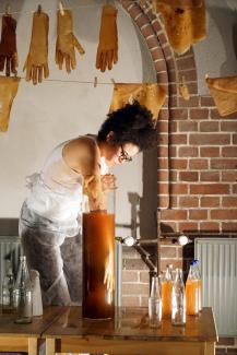 Artist Alanna Lynch in a brick studio space surrounded by fermented glove sculptures hanging on a clothes lines. She is reaching into a tall cylindrical glass vessel full of orange fermented liquid. The vessel of liquid is sitting on a wooden table beside glass bottles, some full some empty. The artist is wearing a thin apron, glasses, and has curly black hair. there is a radiator and an archway in the background, with sunlight filtering through from the right of the picture field.