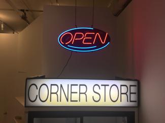 A neon-light 'Open' sign hangs above a drink fridge with a back-lit sign that reads 'CORNER STORE'. Photo taken in Access Gallery's PLOT space.