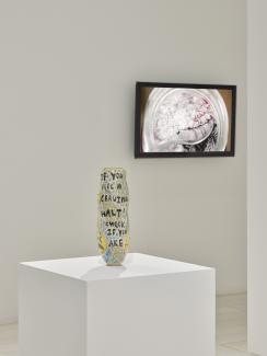 a ceramic vase on a plinth with a video installed on the wall behind, featuring a spoon skimming the surface of a ferment