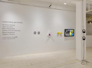 Image of a white wall and a white post in a gallery space. There are texts on the left side of the wall, next to which are two beaded blue and grey plates, a beaded translucent and fuchsia plastic bag, a pair of beaded yellow rubber gloves and a flatscreen displaying the footage of two hands washing dishes in these gloves. There are two sets of black headphones hung on the post to the right of the image.