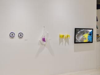 Image of a white wall on which installed two beaded blue and grey plates, a beaded translucent and fuchsia plastic bag, a pair of beaded yellow rubber gloves and a flatscreen displaying the footage of two hands washing dishes in these gloves. 