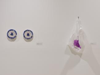 Image of a white wall on which installed two beaded blue and grey plates, and a beaded translucent and fuchsia plastic bag. 