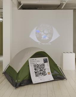 Image of a white gallery space with a video projection showing a large eye and a grey and green tent installation with a large QR code attached to it.
