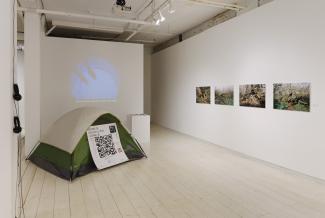 Image of a white gallery space with two perpendicular walls. There is a video projection showing two feet in rain on the end wall, a grey and green tent installation with a large QR code in front of the projection wall, and four colour photograph prints depicting human bodies in nature on the wall to the right.