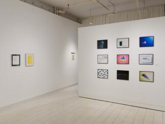 A white gallery space with two perpendicular walls. There are two document-sized prints–one white and one yellow–and a slightly smaller 2D piece on the wall to the left; and nine colour photograph prints of everyday household items displayed in a grid on the wall to the right.