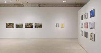 Image of a while gallery space with two perpendicular walls. On one long wall, there are four photograph prints and two document-sized prints. On the side wall to the right, there are nine photograph prints displayed in a grid.