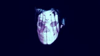 a white face, covered by hands, on which a skull is projected, against a black background