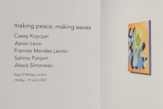 Black vinyl lettering on white wall that reads the exhibition title, artists' and curator's names, and exhibition dates. To the right, there is an angled view of a small brightly coloured abstract painting on a white wall.