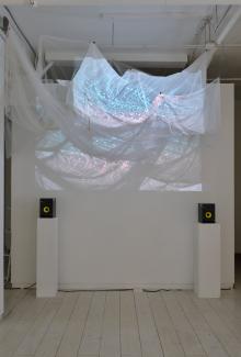 Image of a darkened white gallery space, two black speakers on white plinths flank a fabric installation that hangs above, onto which is projected patterns in blue and red light.