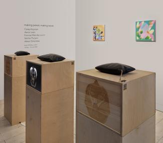 Three wooden plinths on which there sit three light boxes with an image similar to an xray. Atop the boxes are three black vinyl cushions. Behind these plinths there is a white wall with two small, abstract paintings in bright colours.