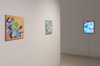A white wall with two small, brightly coloured paintings on the left, on the right, there is a perpendicular white wall in a darkened space on which hangs a brightly coloured lightbox.