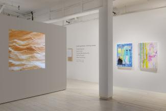 A three-quarter view of the installation. On the left, a square image of carved wood waves is projected on a white wall. On the right, two brightly coloured paintings hang on the wall perpendicular to the projection.