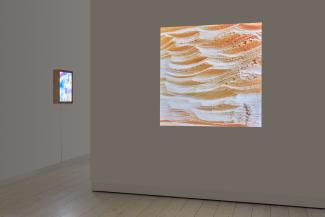 A three-quarter view of the installation. On the left, the is a brightly coloured lightbox. On the right wall, perpendicular and in front of the lightbox, a square image of carved wood waves is projected on a darkened white wall. 